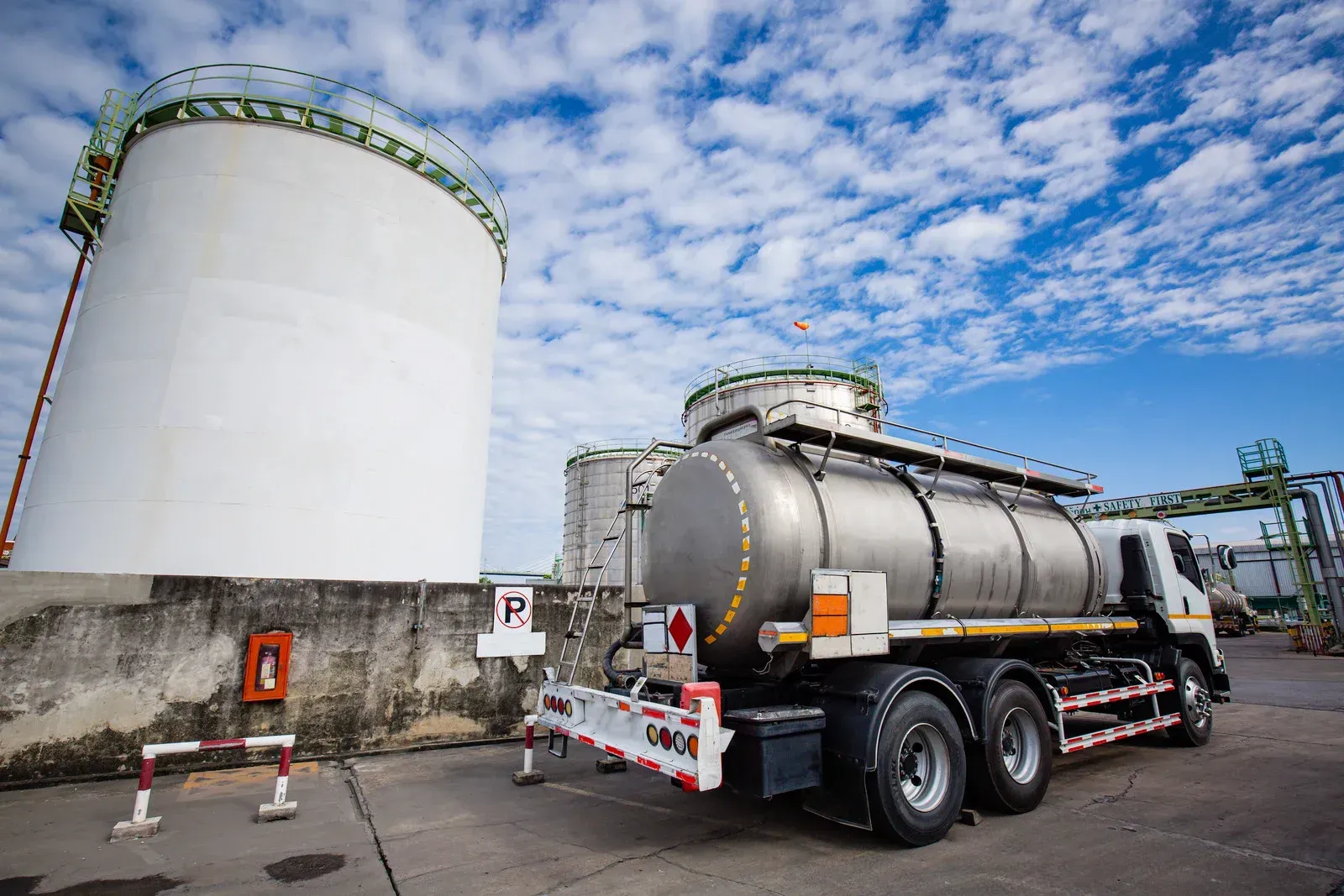 A tanker truck parked next to a white storage tank at a fuel depot on a sunny day.