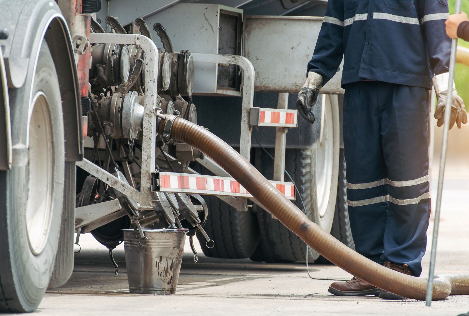 A sanitation worker in navy blue uniform unloads waste from a truck using a large hose into a metal bucket on the pavement.