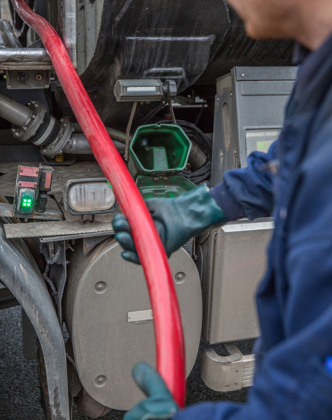 Person in blue overalls and gloves connecting a red hose to a tanker truck for refueling.
