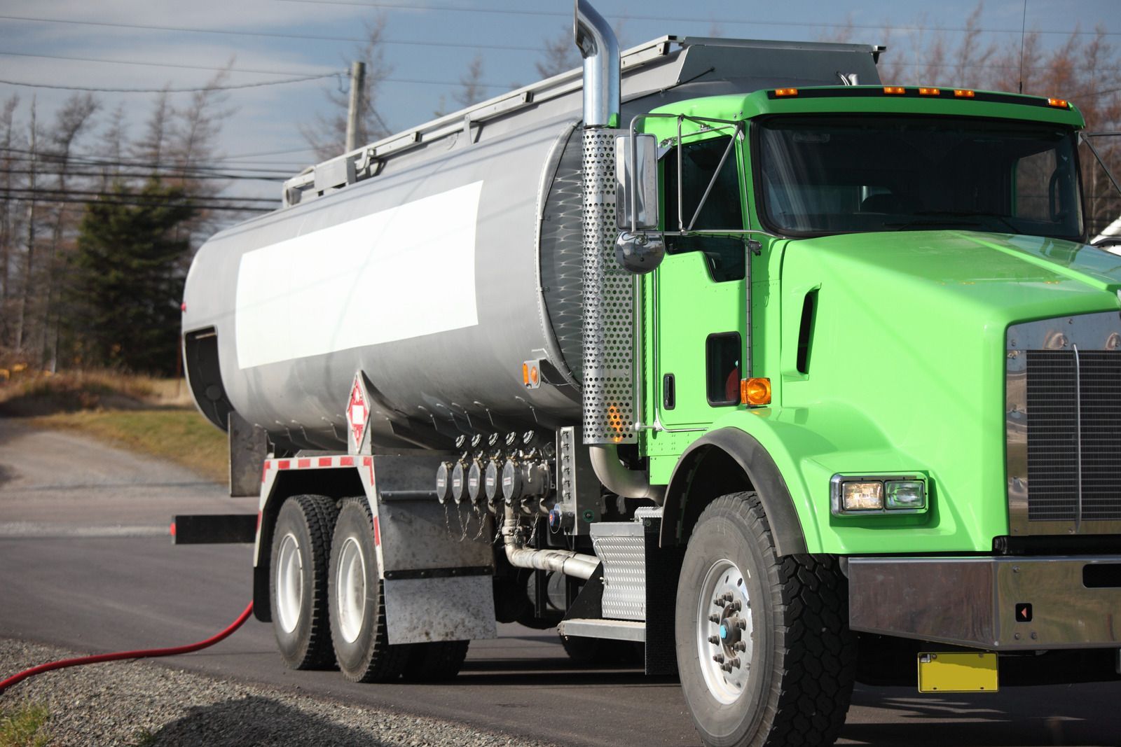 A green tanker truck on a road, with a silver tank. A red hose is connected to the truck.