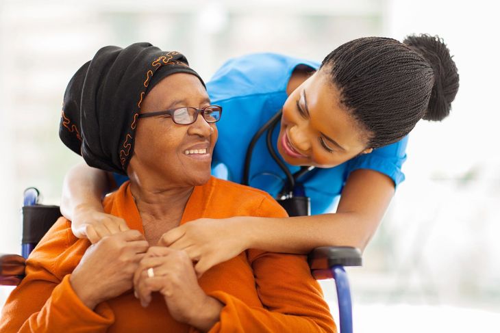 Nurse in blue scrubs comforting an elderly Black woman in a wheelchair; both are smiling.