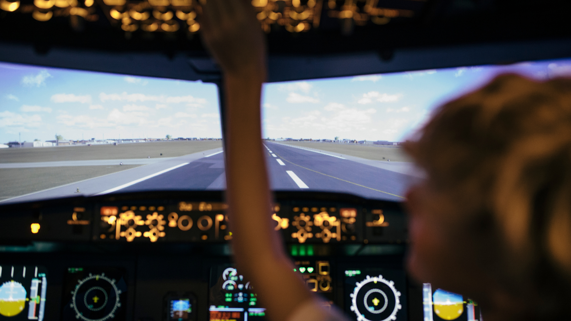 Person in a flight simulator cockpit, reaching towards controls with runway in background.