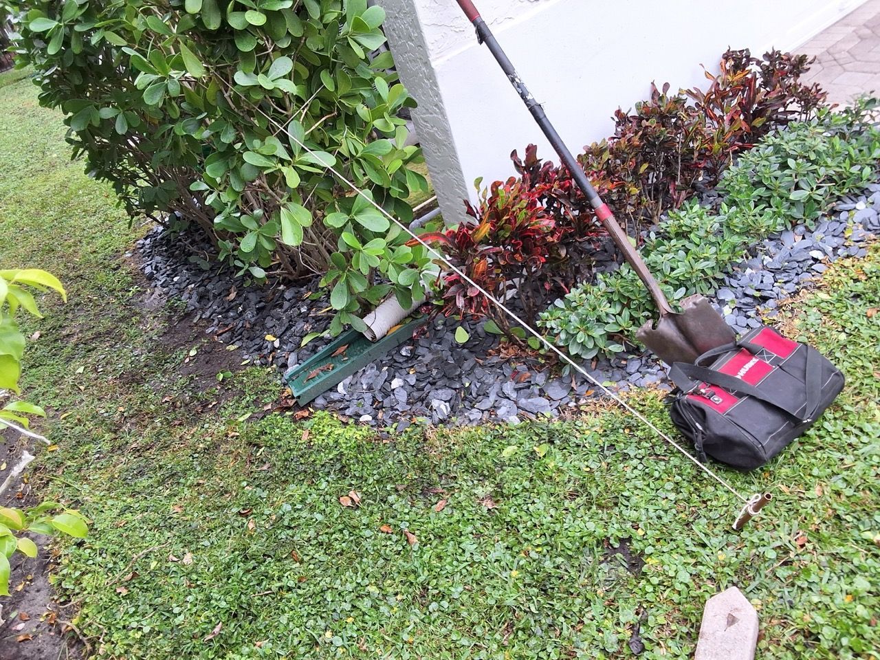 Gardening tools on grass beside a brick wall and shrubs, with a red-and-black bag and rake.