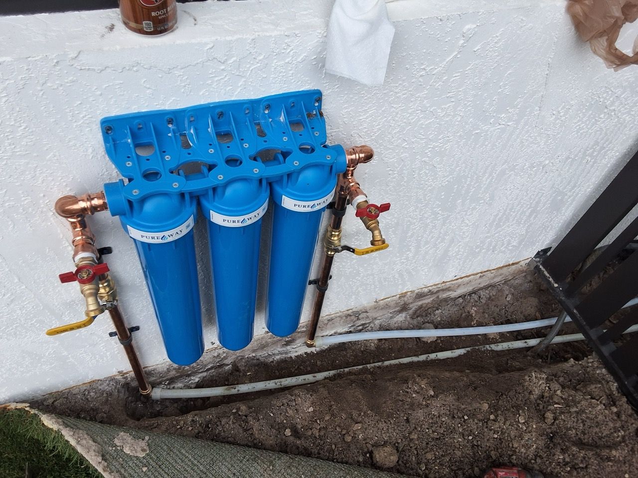 Blue water filtration system with four canisters mounted on an outdoor wall and connected to pipes and valves