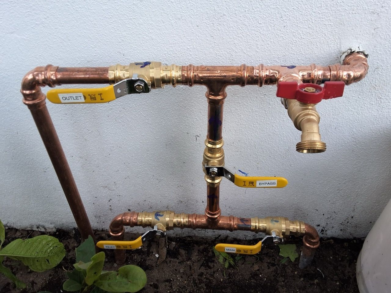 Outdoor copper plumbing pipes with yellow and red valves against a wall, near plants