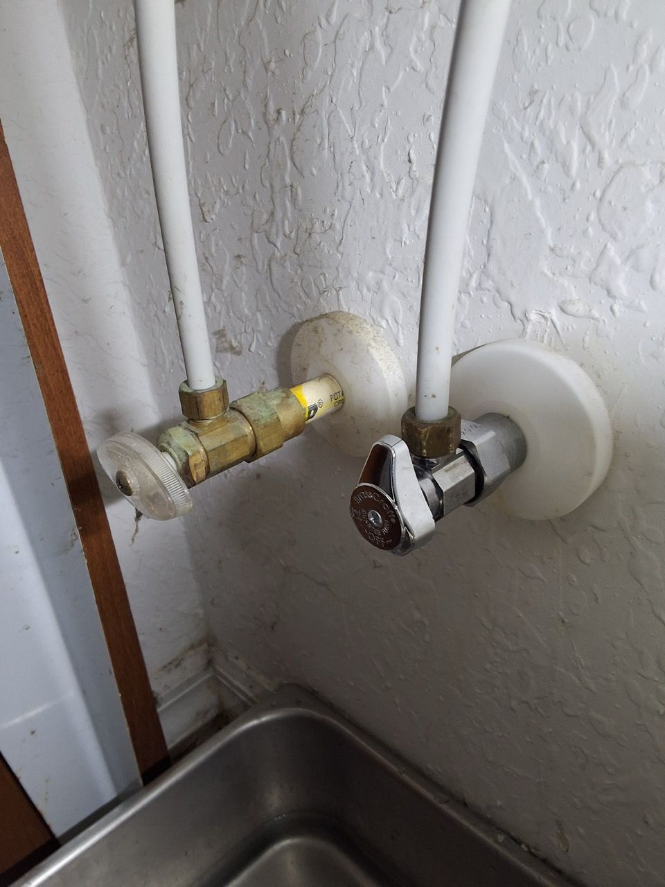 Close-up of two wall-mounted water shutoff valves and white pipes above a stainless steel sink.