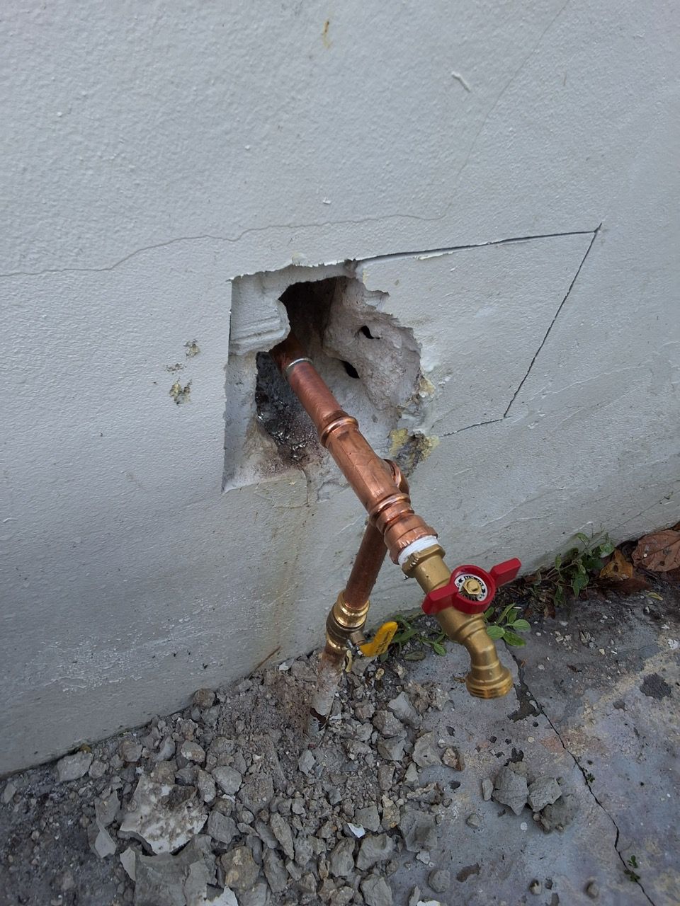 Copper and brass outdoor water pipes protruding from a hole in a concrete wall, with rubble below