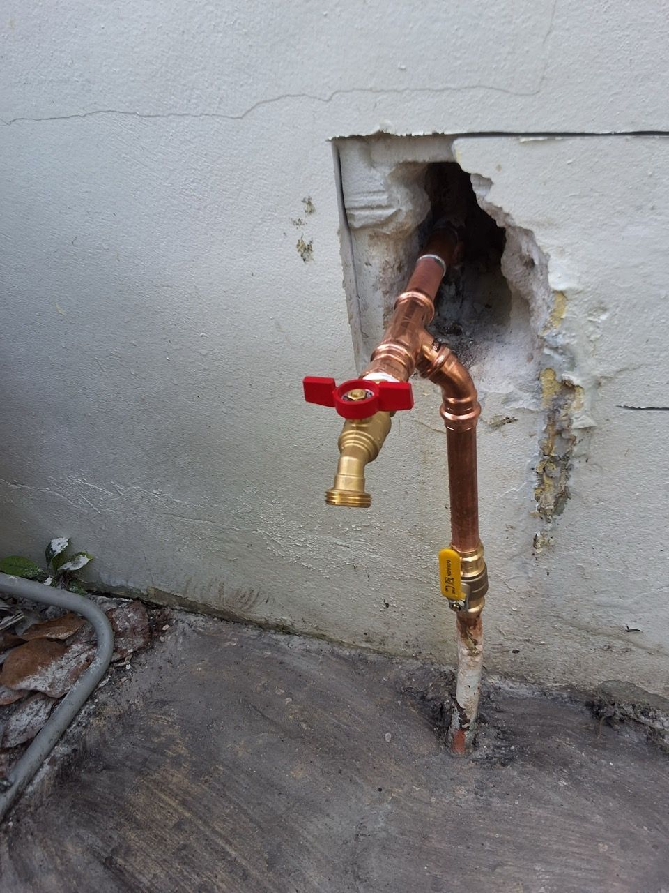 Red valve and copper pipes protruding from a hole in a gray exterior wall.
