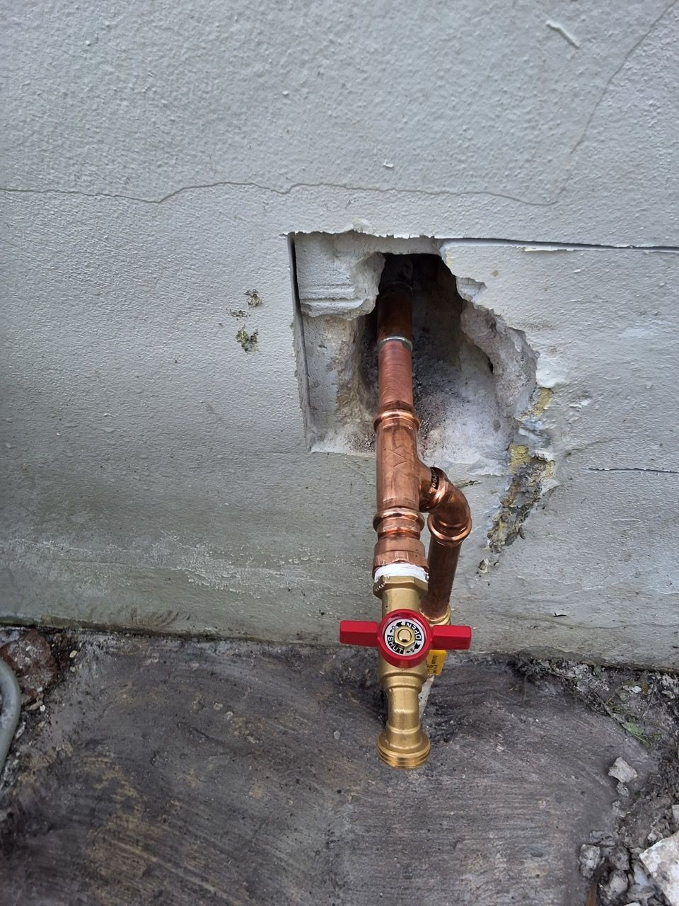 Exposed copper pipe with red valve sticking out of a hole in a gray concrete wall.