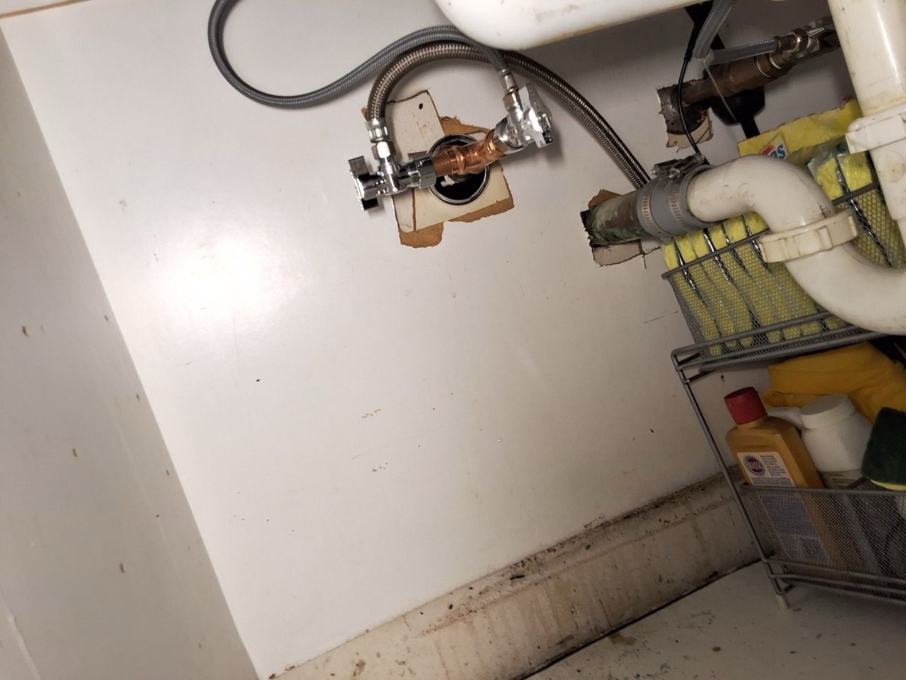 Under-sink plumbing with pipes, valves, and a water shutoff assembly against a white wall.