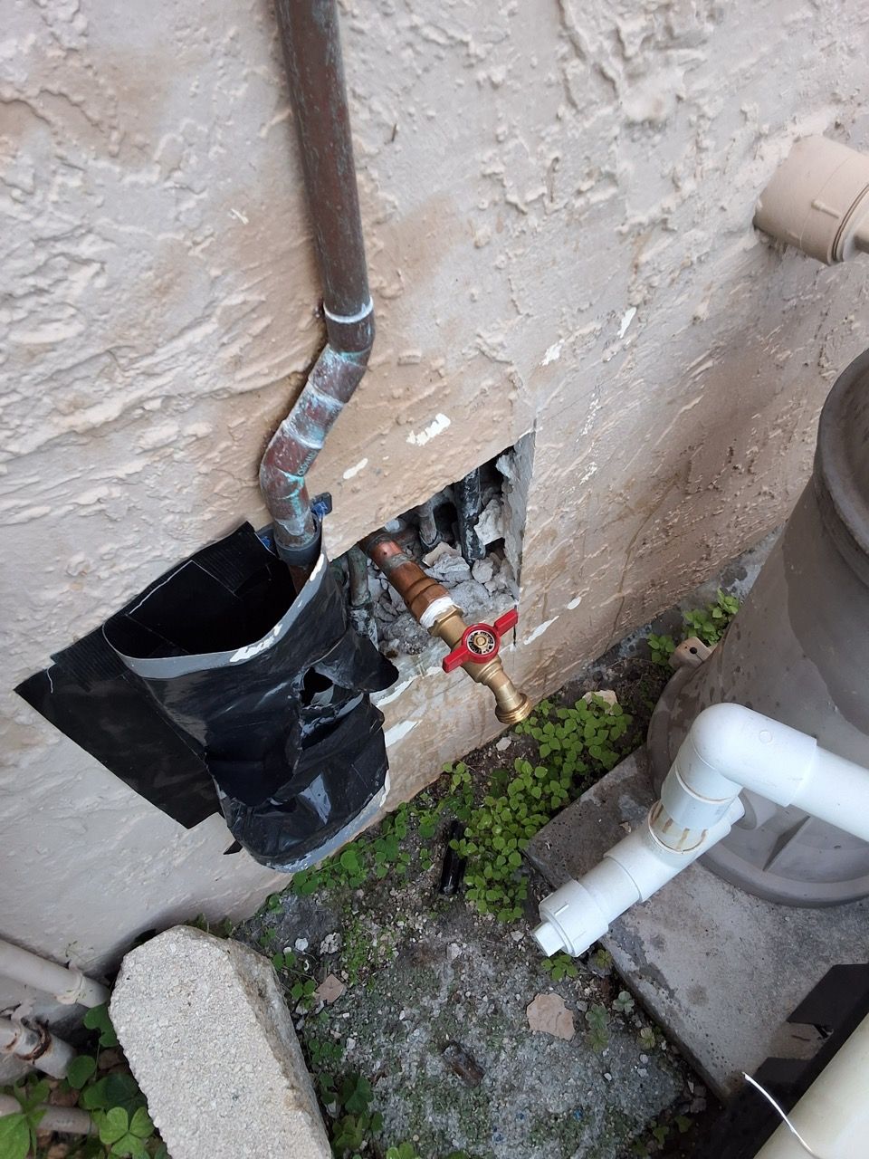 Broken wall vent with exposed black ductwork and plumbing pipes outside a building