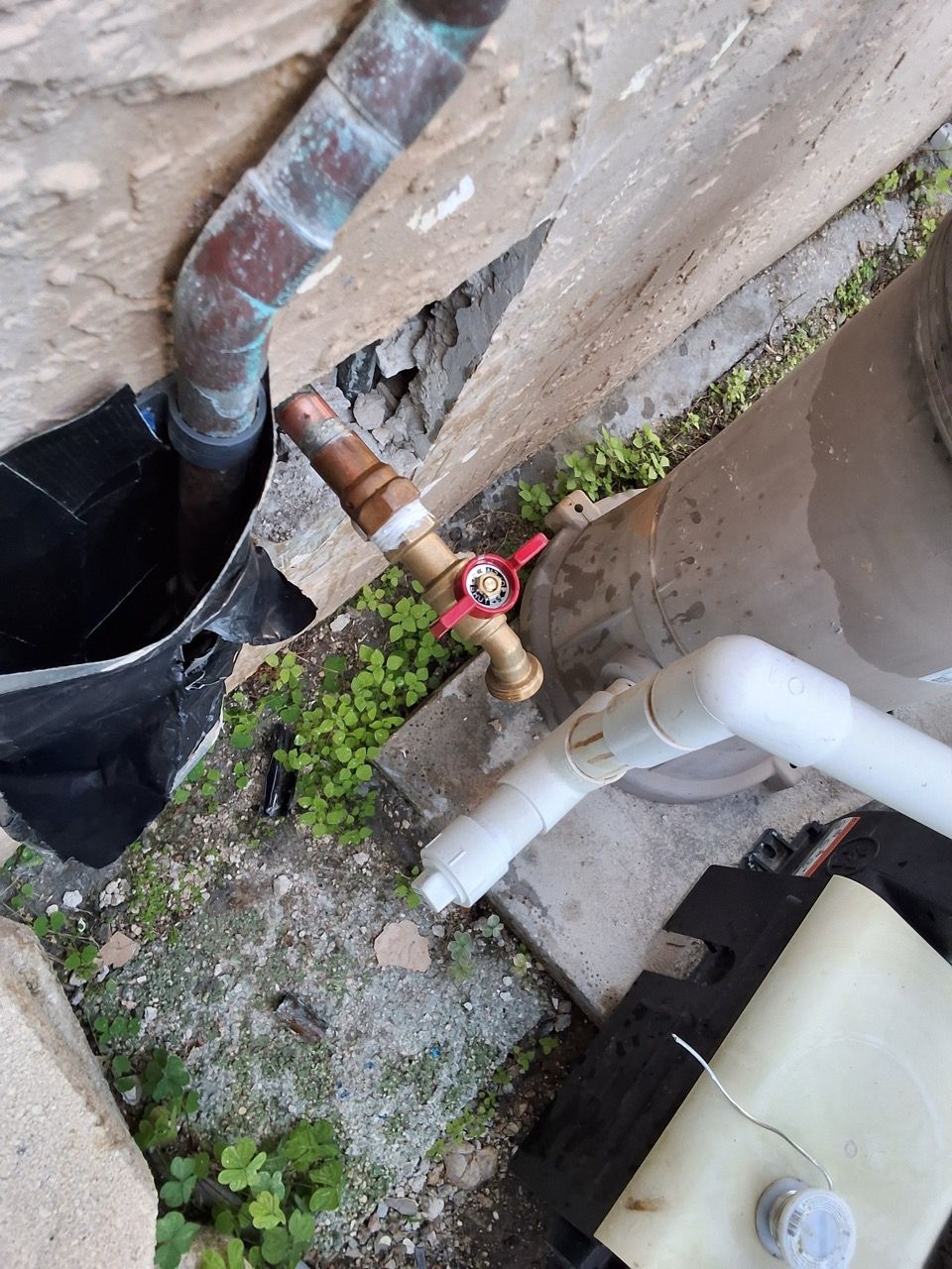 Outdoor plumbing pipes and a red shutoff valve beside a wall, with weeds and a black drain basin.