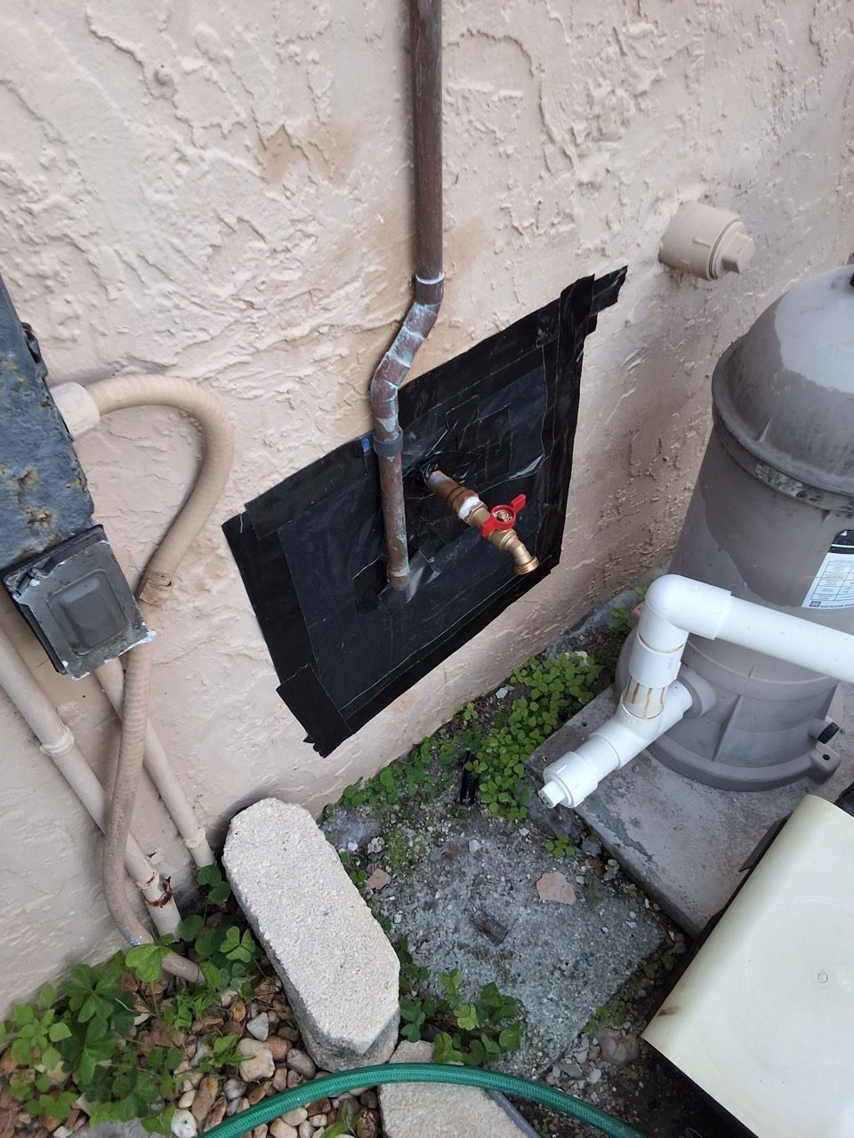 Outdoor wall with exposed pipes, a black access panel, and a gas meter beside gravel and a hose.