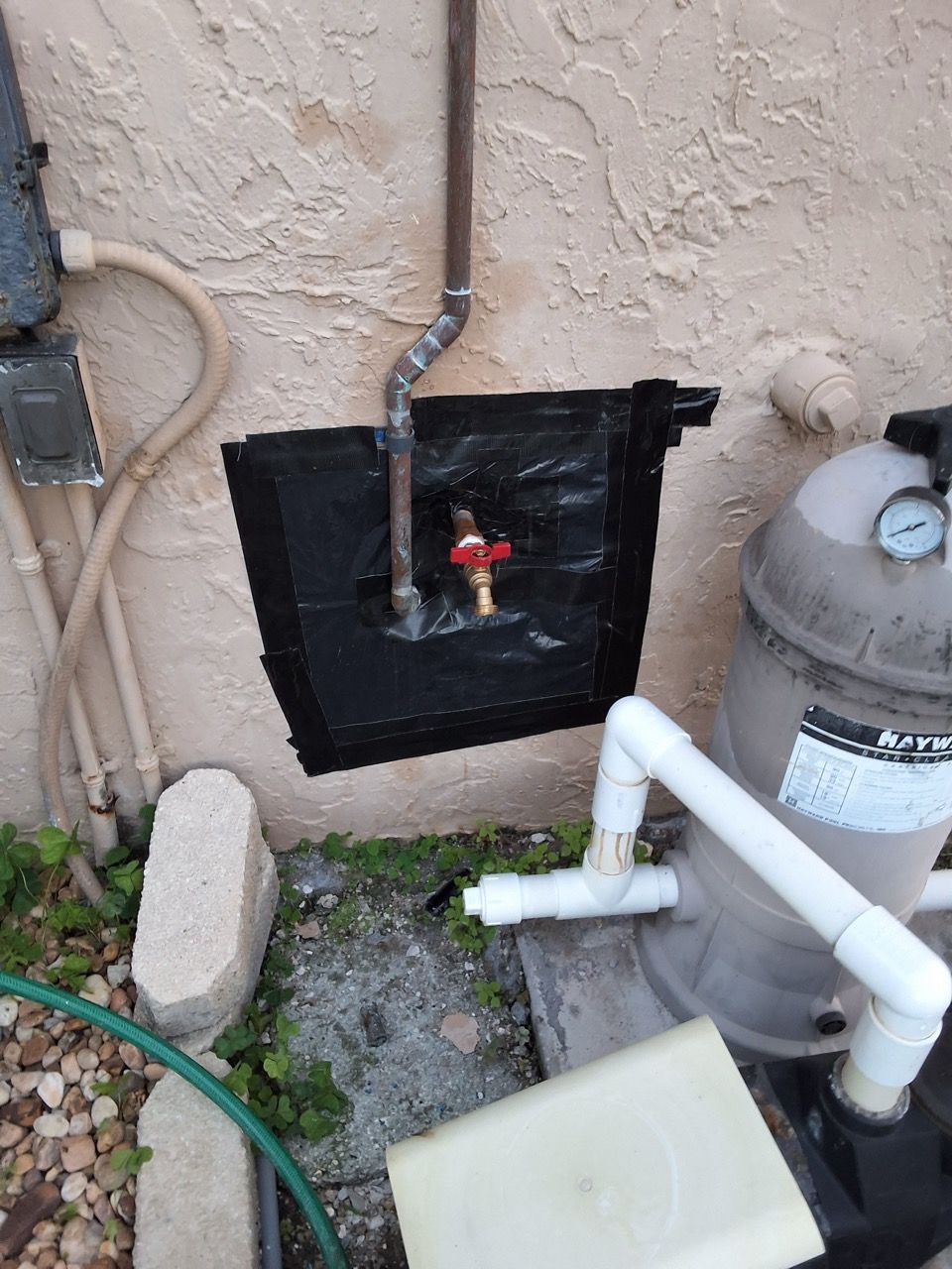 Outdoor utility area with exposed pipes, red shutoff valve, and a gray gas meter against a stucco wall
