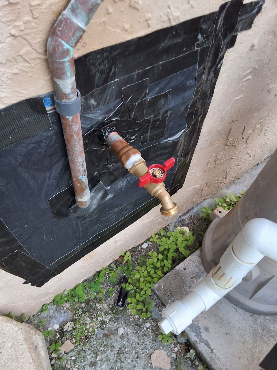 Outdoor plumbing area with corroded pipe, brass shutoff valve, and white PVC drain against a wall