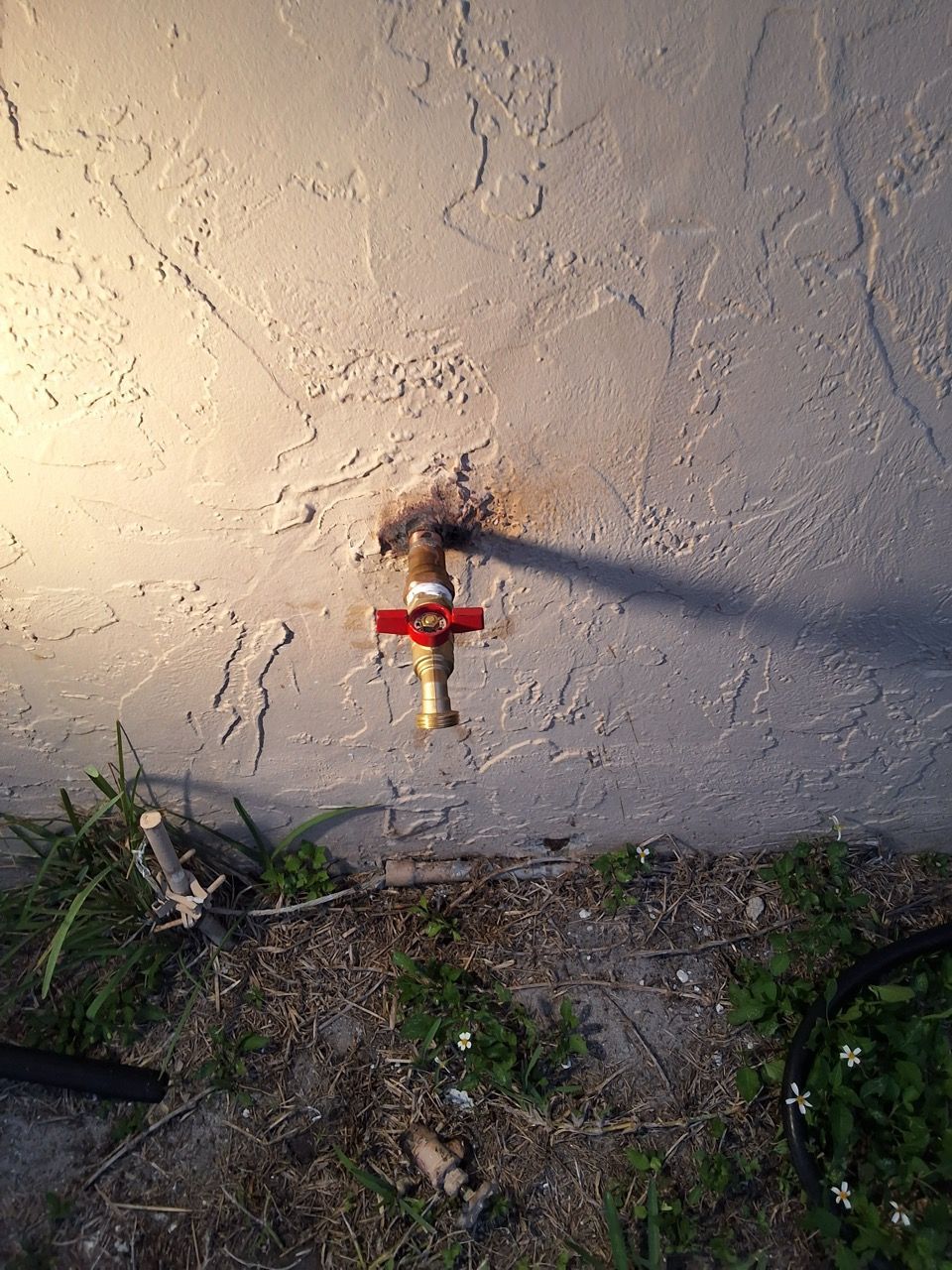 Red water spigot mounted on a stucco wall beside patchy grass and dirt.