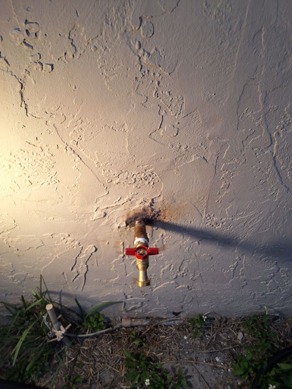 Outdoor wall with a red water valve and brass pipe near the ground, casting a long shadow.