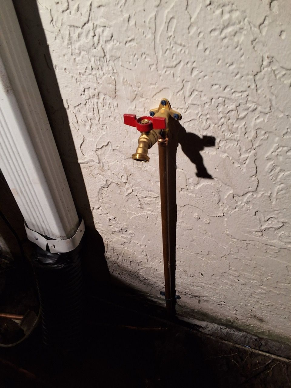 Outdoor faucet with a red valve on a wall beside a white pipe and dark ground.