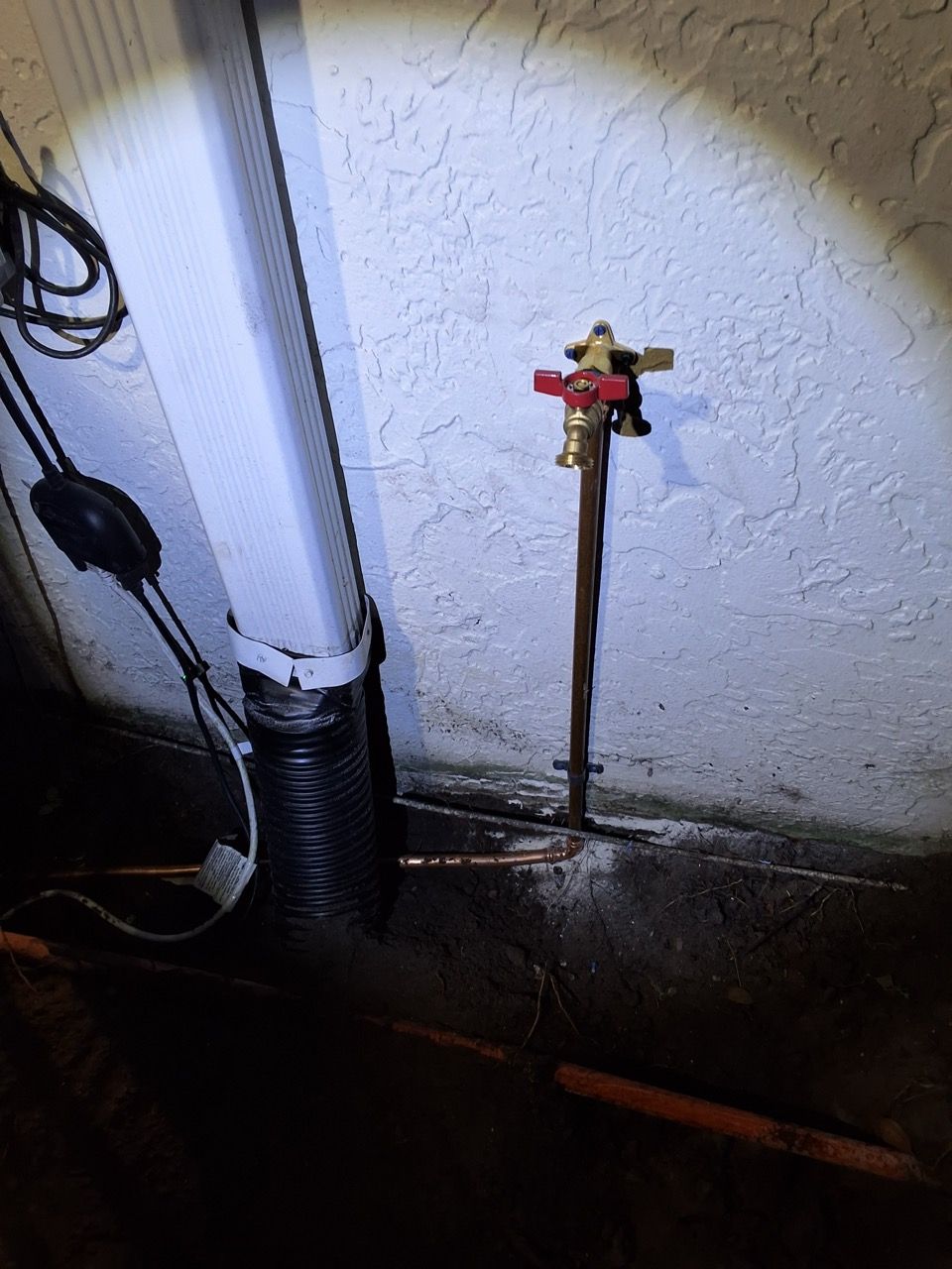 Red outdoor water spigot mounted on a stucco wall beside white and black pipes at night
