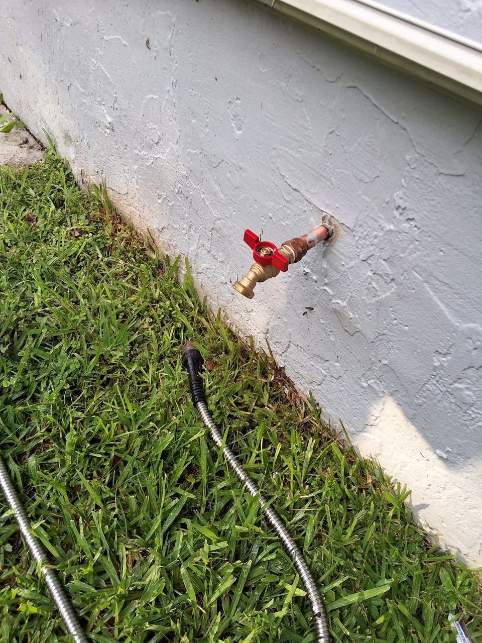 Red outdoor water faucet on a house wall above grass, with a hose nearby