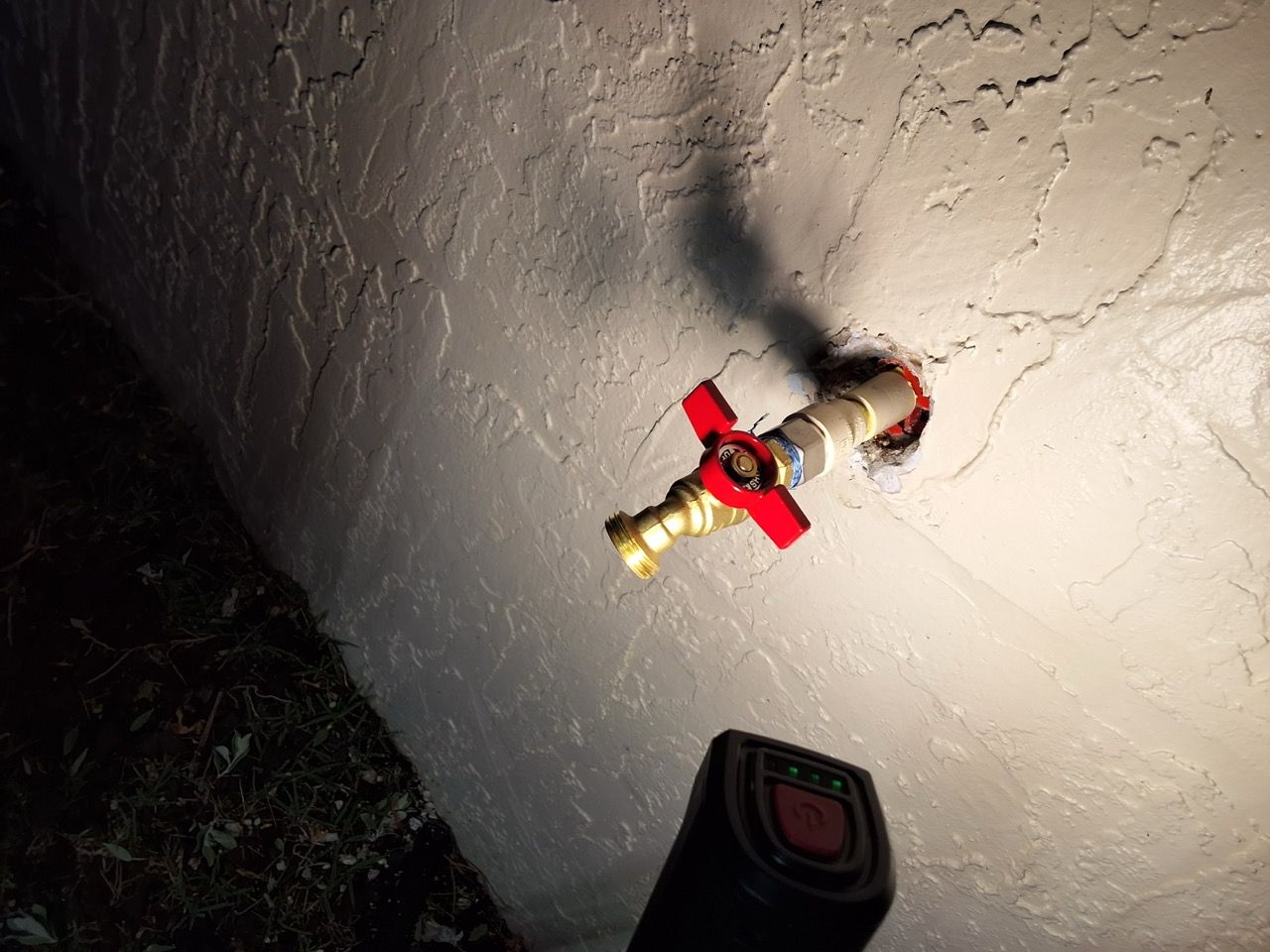 Red-handled outdoor water faucet on a wall, lit by a flashlight at night.