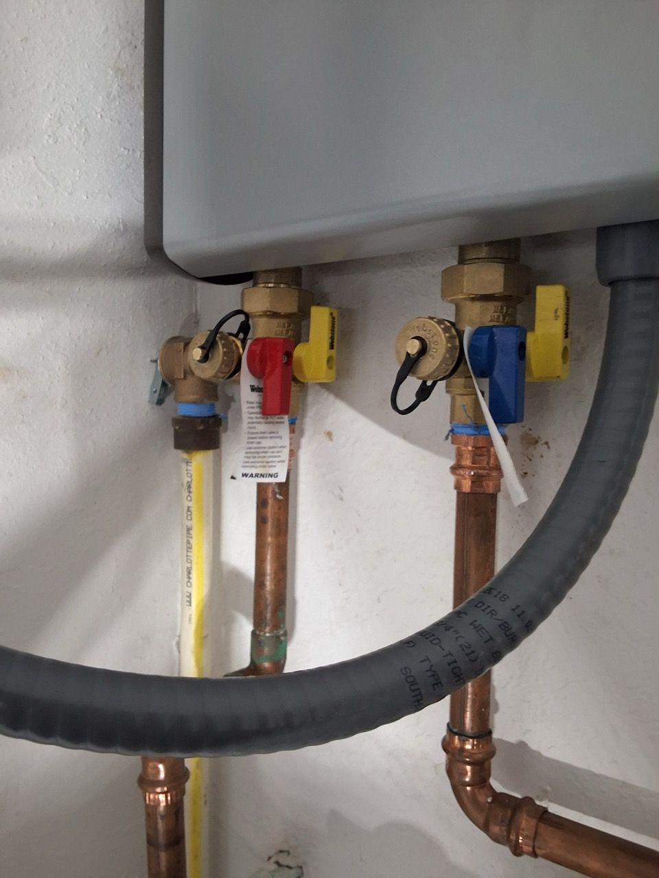 Plumbing pipes and shutoff valves beneath a sink, with yellow and red tags and a black hose in front.
