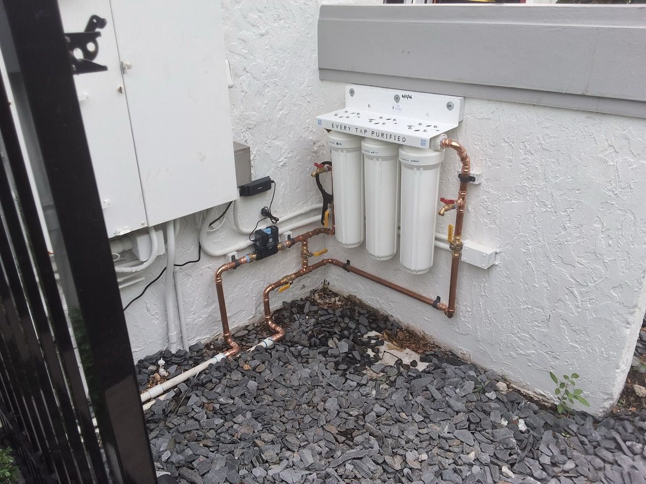 Outdoor utility pipes and filters mounted on a gray wall beside gravel ground