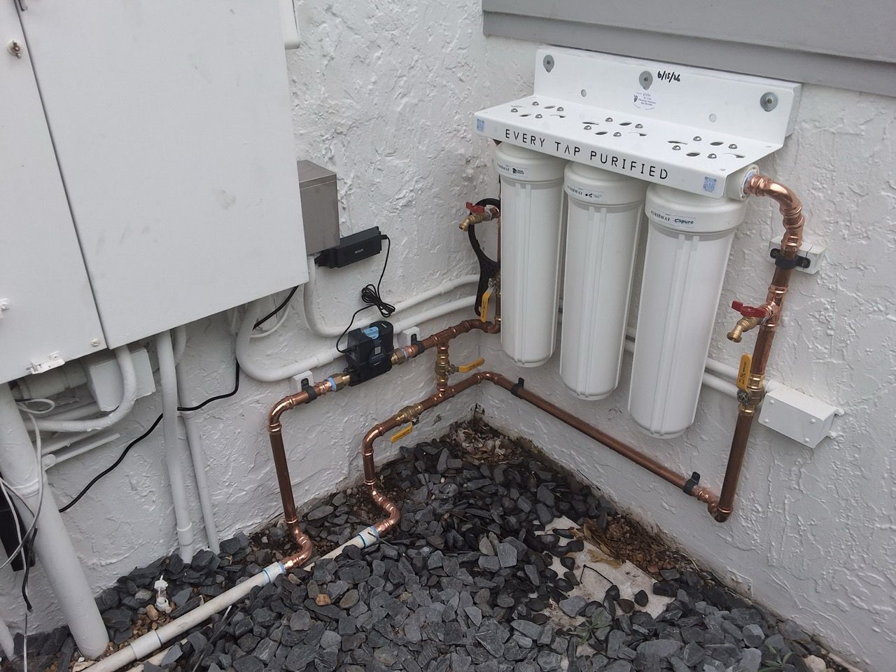Outdoor water filtration system with three white filter canisters, copper pipes, and a gravel base against a wall