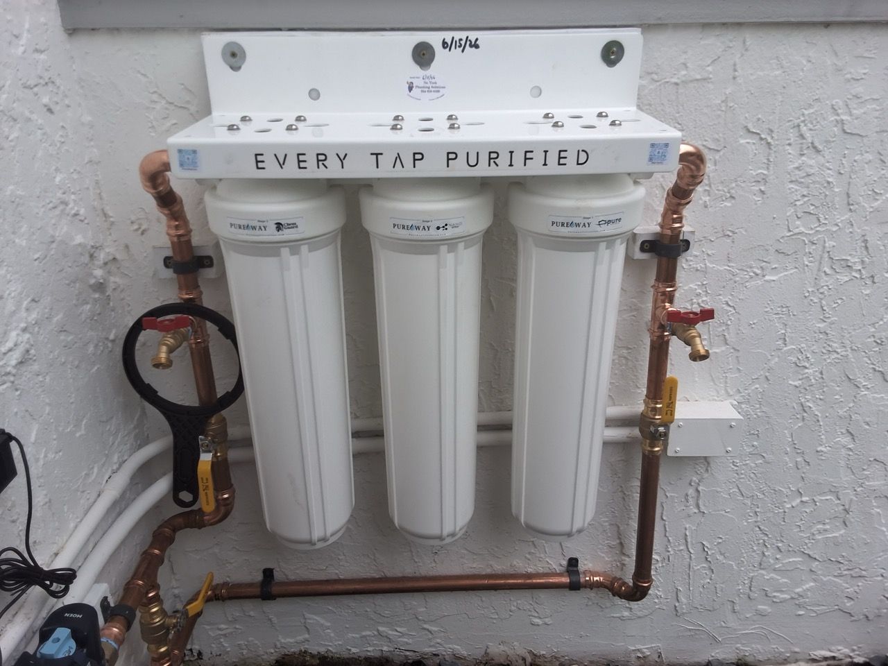 Wall-mounted three-cartridge water filter system with copper pipes and a white tank, labeled “Every Tap Purified.”