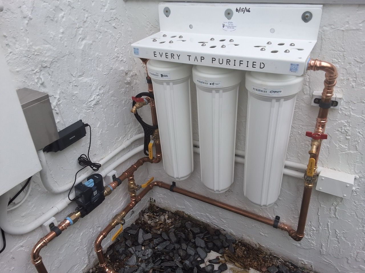 Wall-mounted water filtration system with white filters and copper pipes beside a small gravel pit
