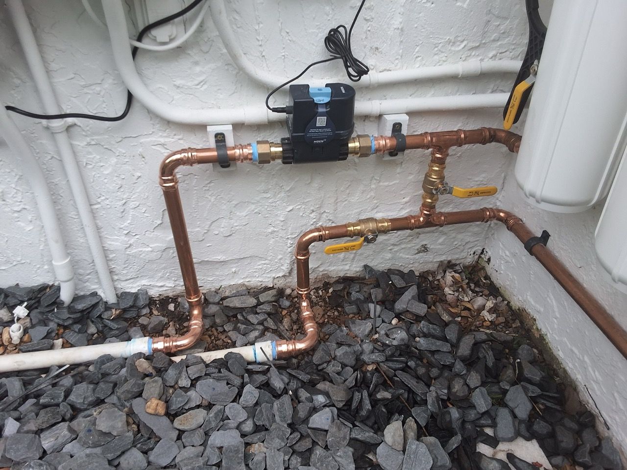 Copper pipes and valves on a white exterior wall beside a black utility meter and gravel bed.