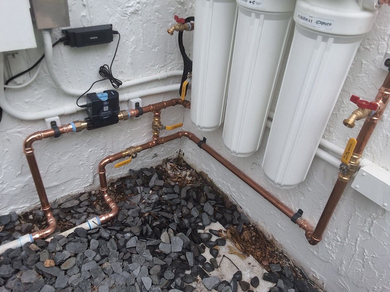 Under-sink water filtration system with white canisters, copper piping, and a black drain tray over gravel.