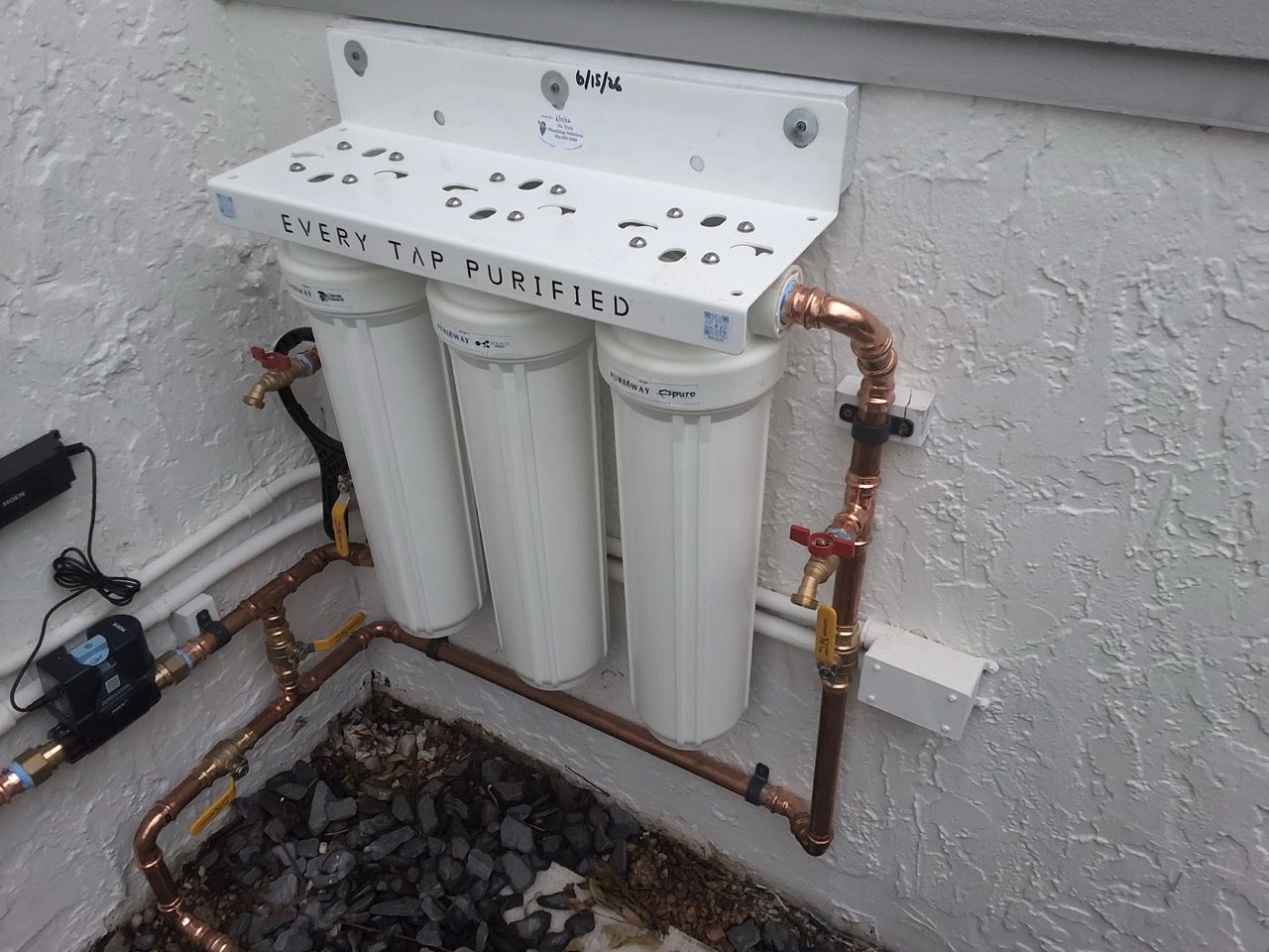 Wall-mounted three-canister water filter system with copper piping above a gravel-lined pit.