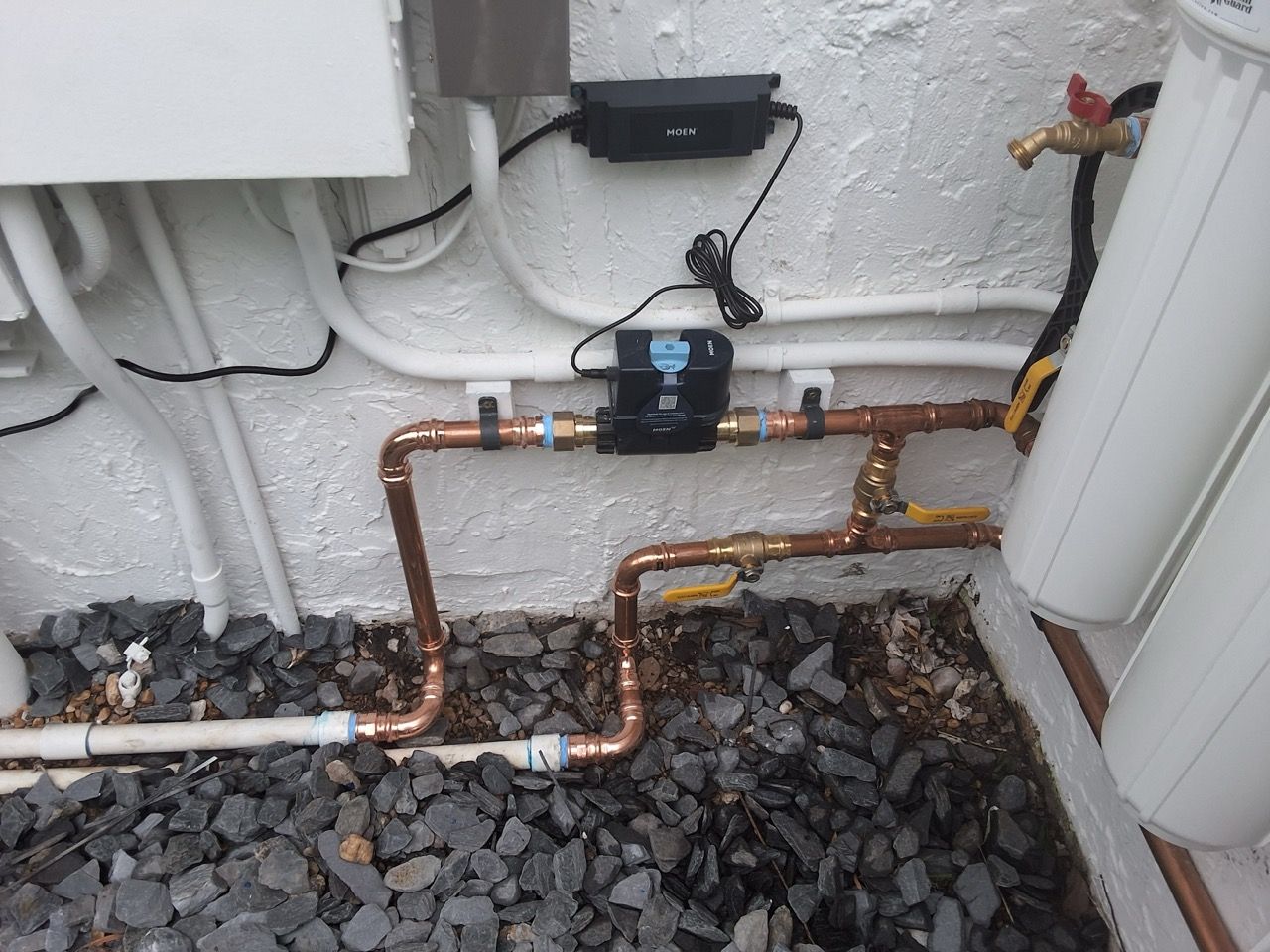 Copper plumbing pipes and a black valve beneath a white exterior wall, surrounded by gravel.