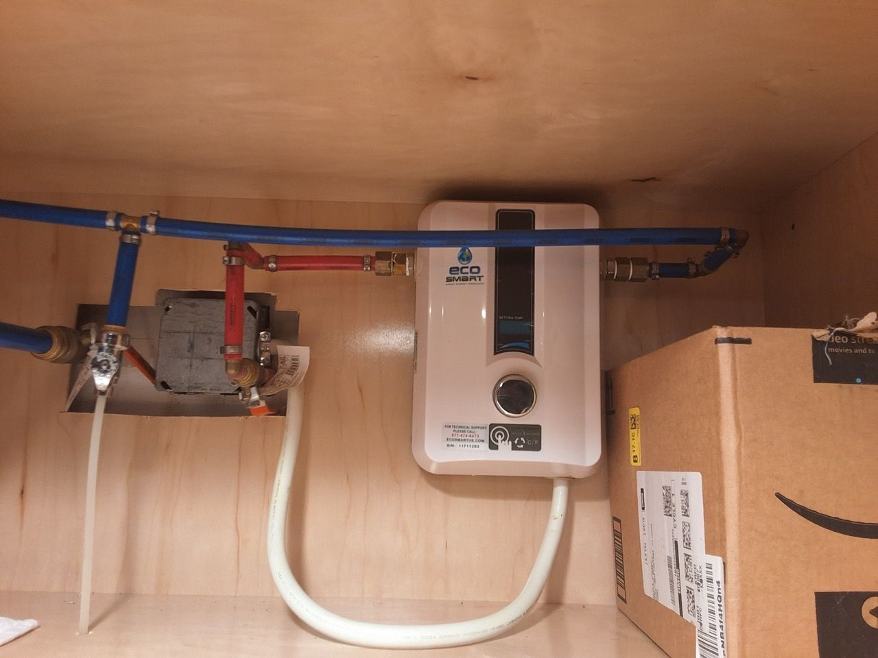 Under-sink water heater with pipes, valves, and white hoses inside a wooden cabinet
