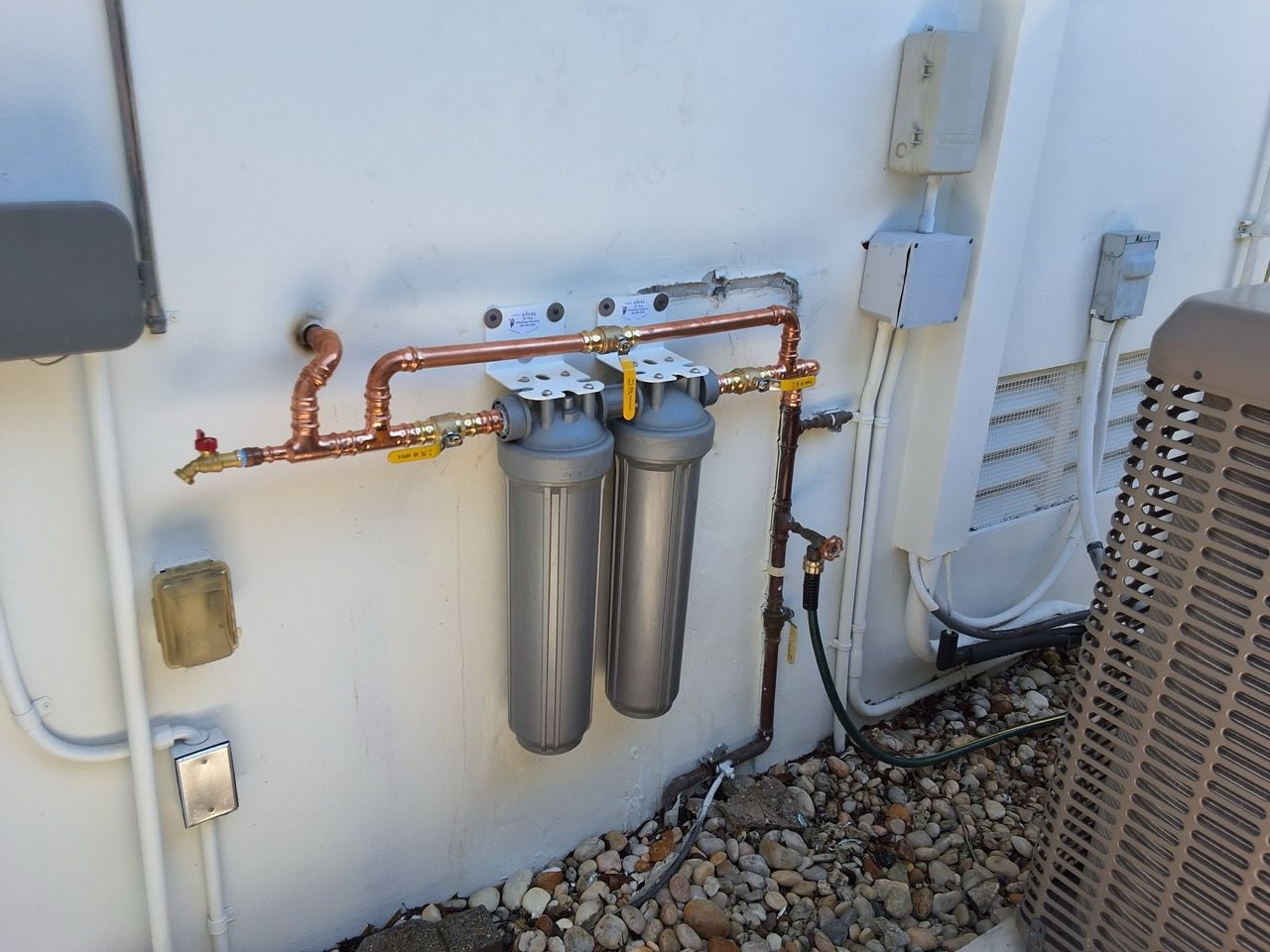Wall-mounted water filtration system with copper pipes, twin metal canisters, and electrical boxes outdoors.