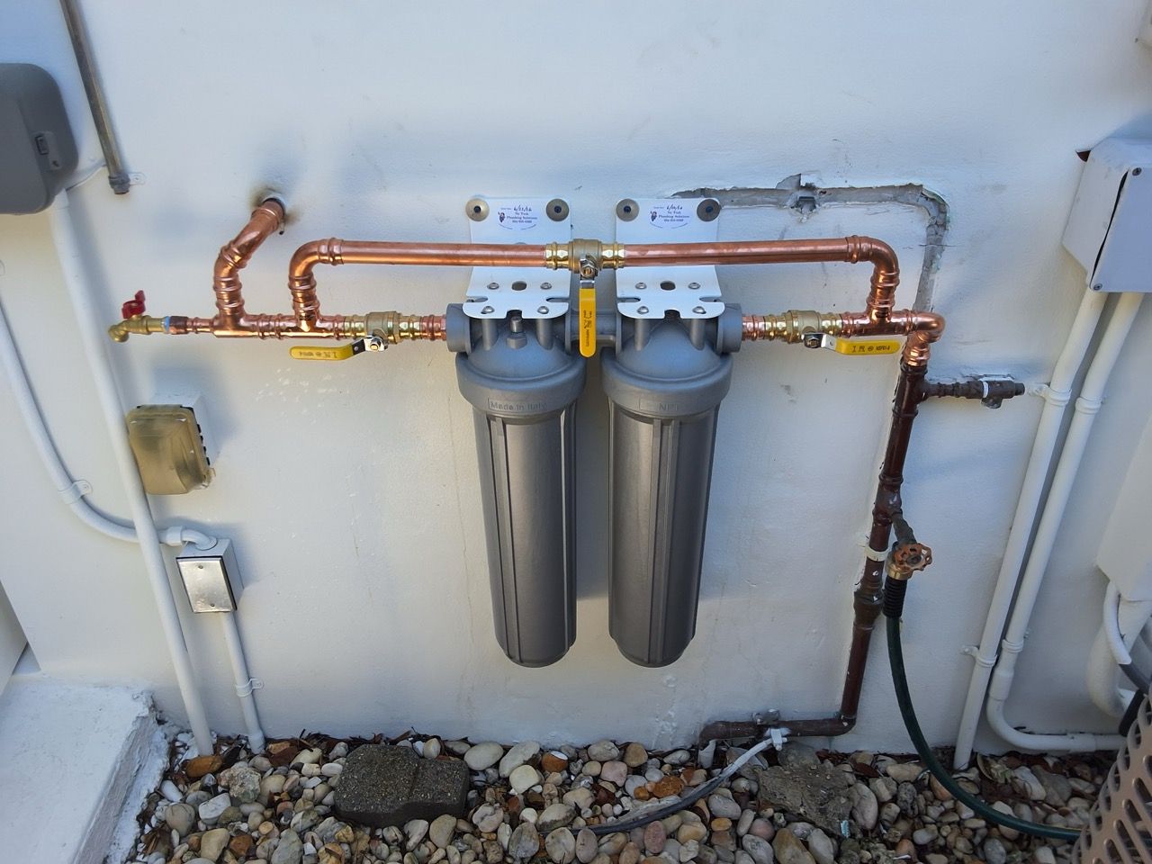 Two gray water filters mounted on a wall with copper piping and shutoff valves beneath them