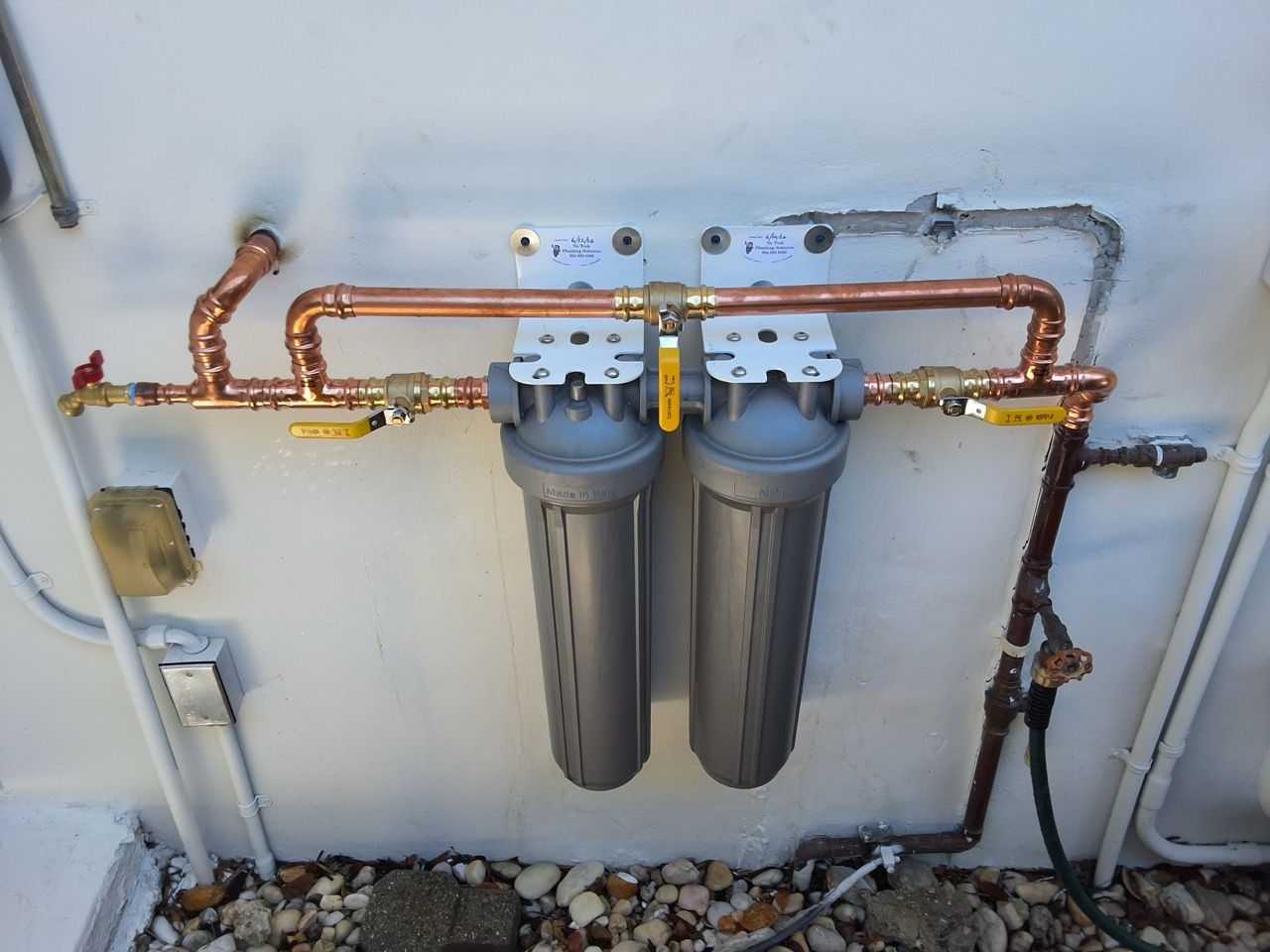 Two-cylinder water filtration system with copper piping mounted on a white wall outdoors