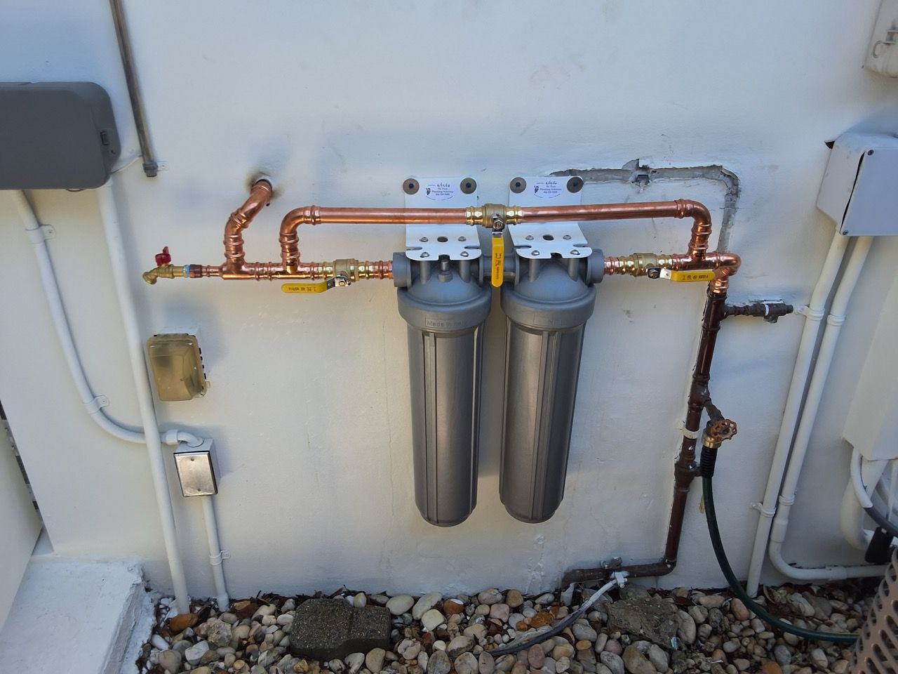 Two gray water filter housings with copper pipes mounted on a white exterior wall
