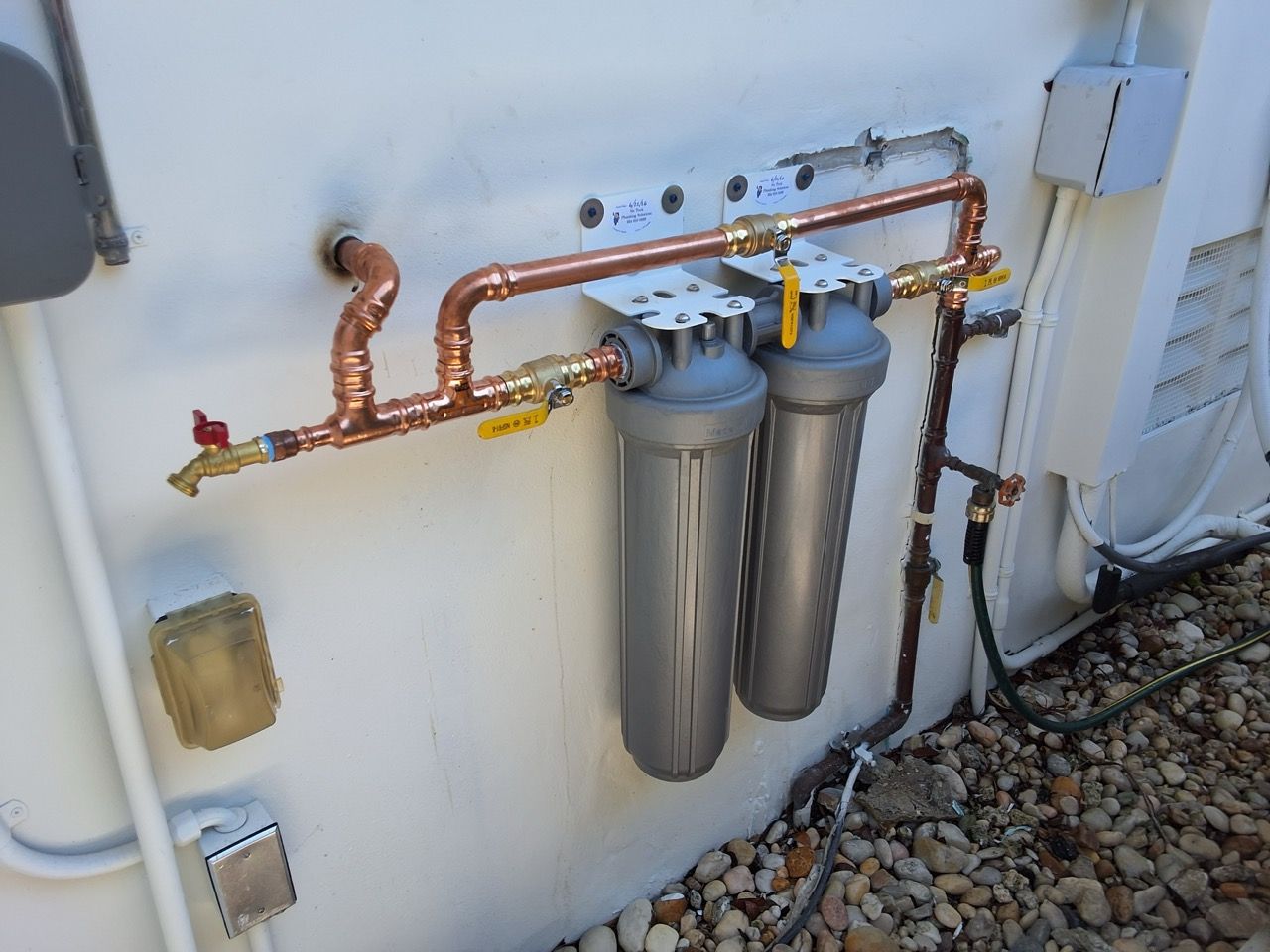 Exterior wall-mounted water filtration system with copper pipes and two large filters next to gravel ground