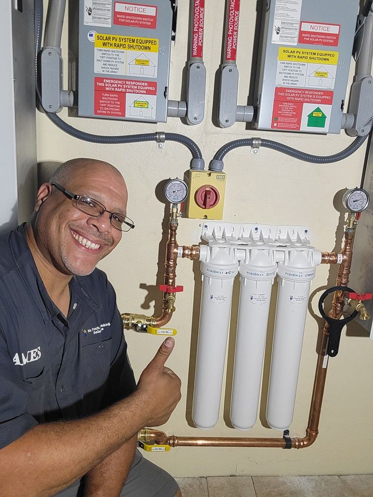 Smiling man in navy shirt kneels beside a water filtration system with pipes and filters.