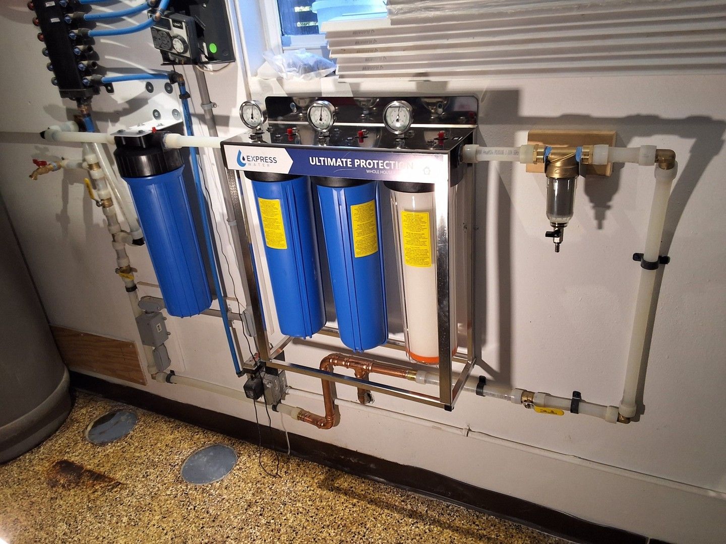 Water treatment filter system with blue canisters, pipes, and valves mounted along a wall.