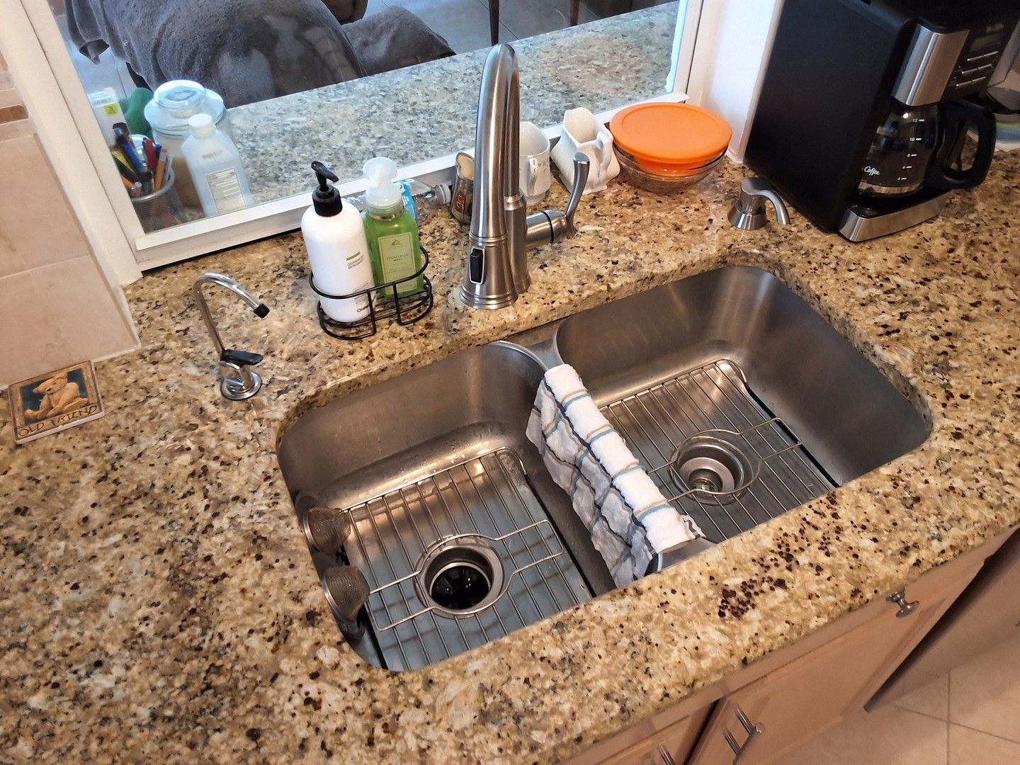 Double kitchen sink in speckled countertop with soap and cleaning items by the faucet.
