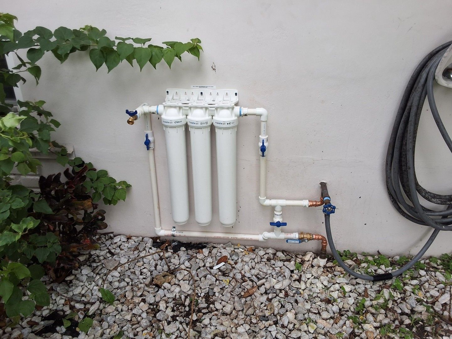 Water filtration system with four white canisters mounted on a wall beside a hose and gravel ground.