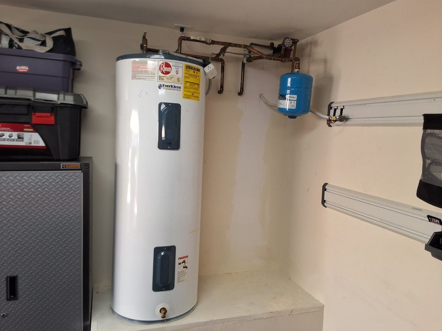 Water heater in a garage corner with shelving and a blue expansion tank above it.