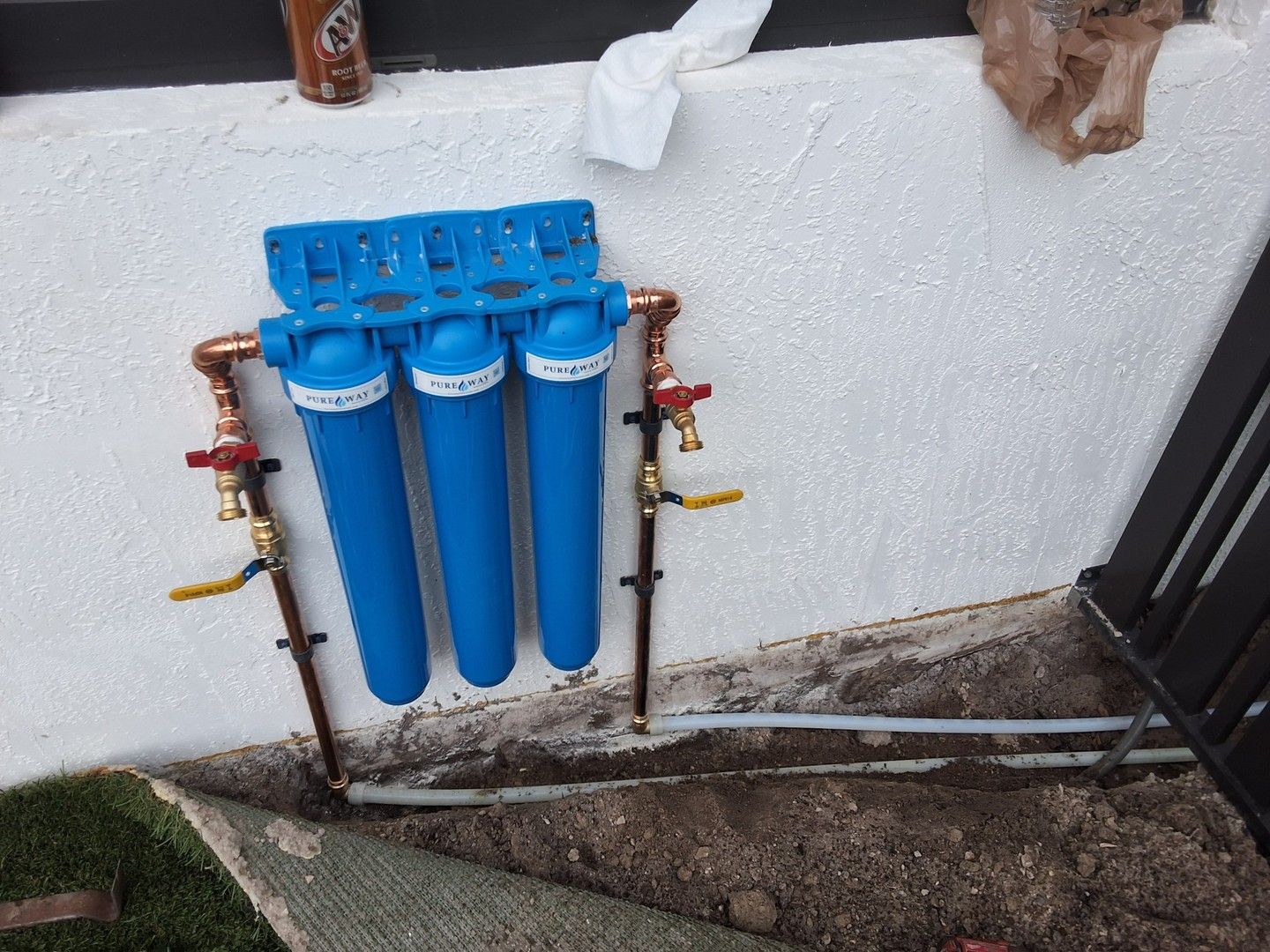 Blue water filtration system with three filters mounted on an outdoor wall beside pipes and grass