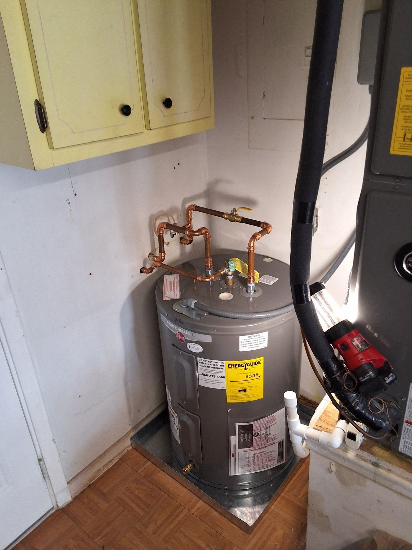 Water heater in a utility closet beside yellow cabinets, with copper pipes and a black vacuum nearby.