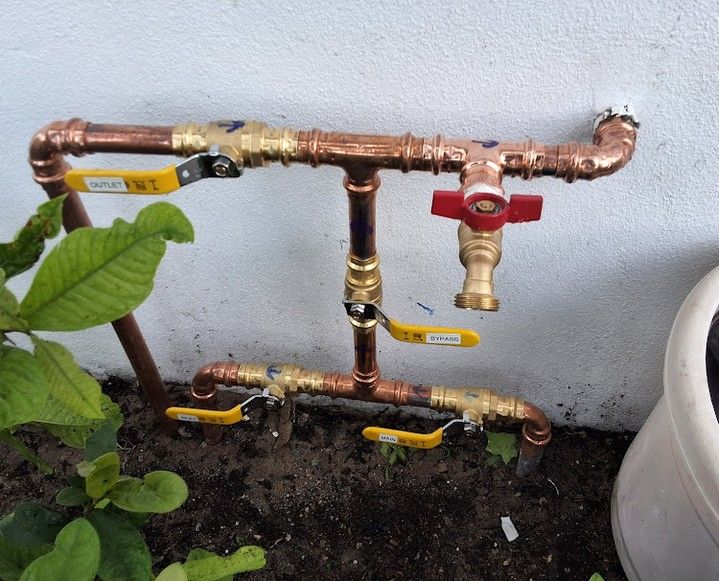 Copper outdoor pipe assembly with yellow valves and a red shutoff valve above garden soil