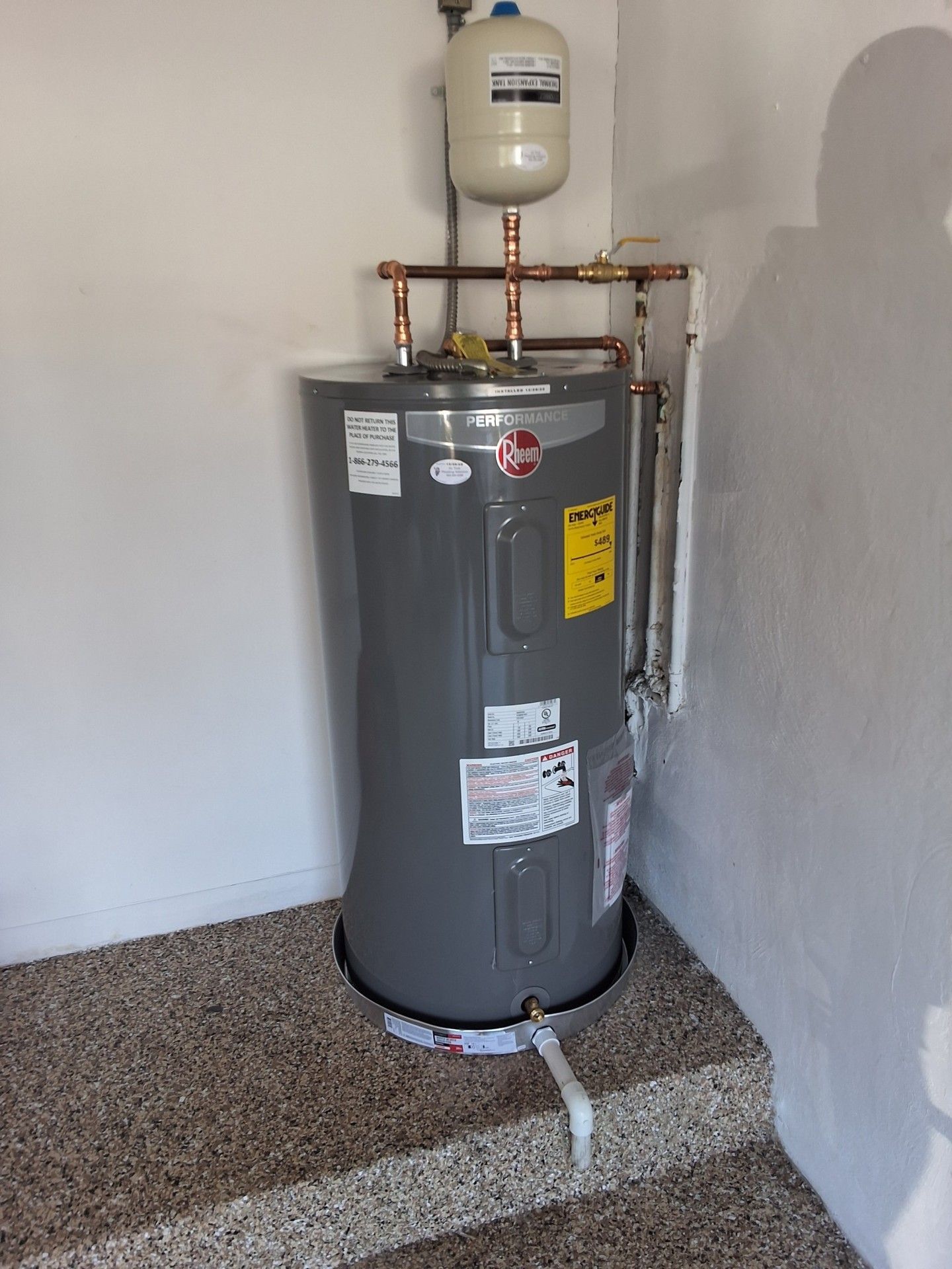 Water heater in a corner on a tiled platform, with pipes and a small expansion tank above it.