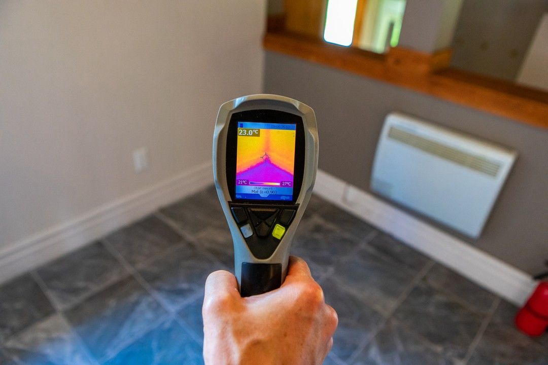 Handheld thermal camera aimed at a room corner, showing heat map of wall and floor.